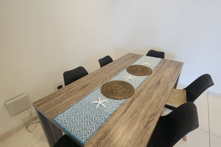 Wooden table with six black chairs, placemats, and a blue runner in a minimal dining room.