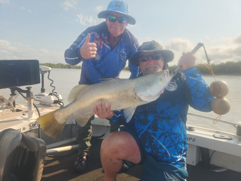 Two men on a boat holding a large fish, both wearing blue shirts and sun hats.