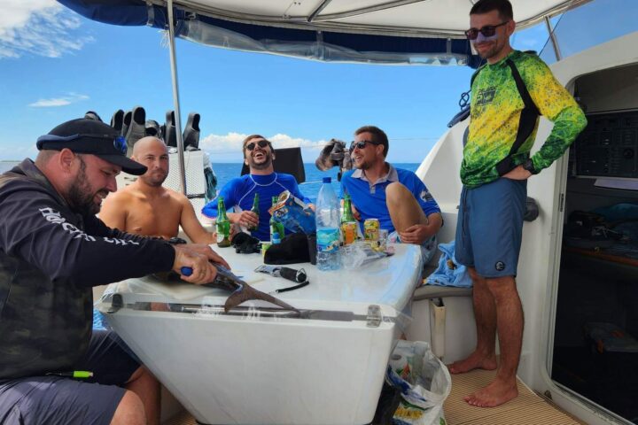 Five men on a boat, one cleaning a fish, others smiling around a table.