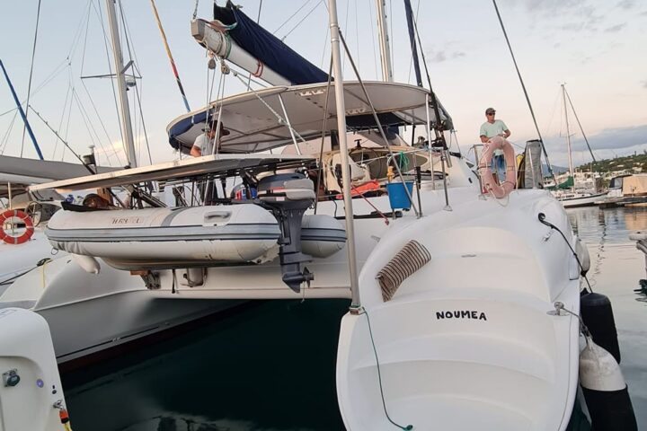 Docked catamaran with sails down, small motorboat, and people onboard.