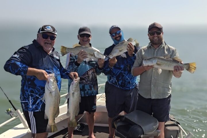 Four men on a boat holding large fish, smiling with ocean in the background.